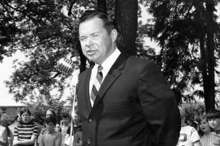 Black and white photo of a man in a suit and tie speaking at a microphone